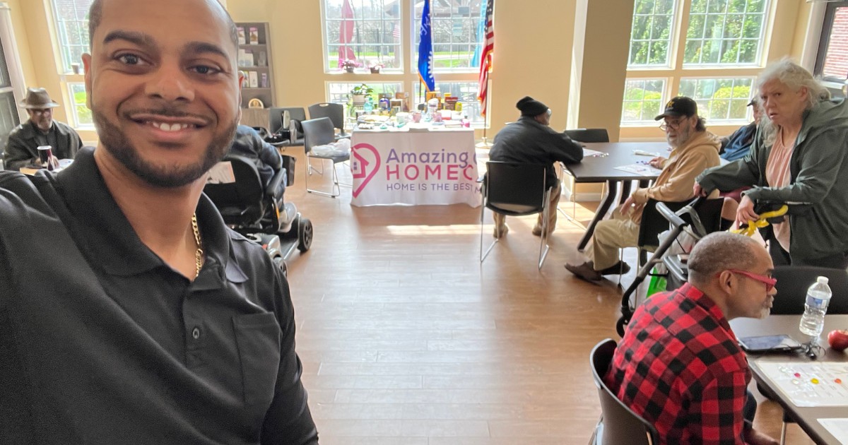 A selfie photo taken by a smiling Black man in a black polo shirt, capturing a sunlit community room at Lyons Place 2 with seniors and veterans playing games at tables. In the background, an Amazing Grace Homecare table with branded cloth and American and military flags is central. Several seniors use wheelchairs or walkers. The mood is cheerful.
