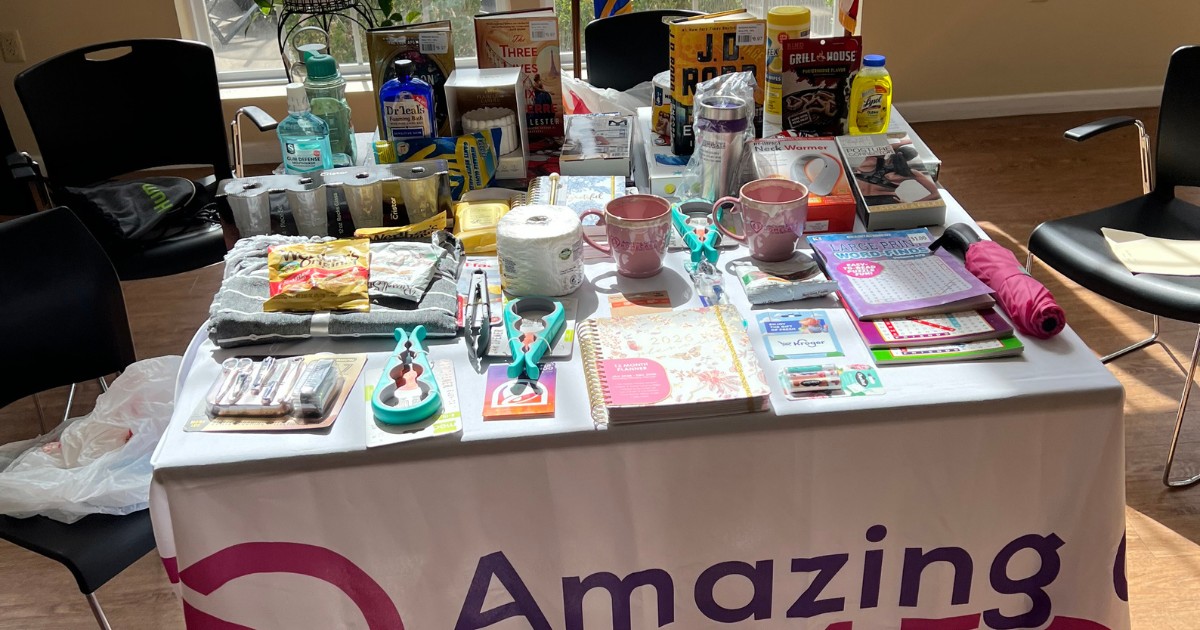 A detailed close-up photograph of a richly stocked prize table at an event, covered in books, mugs, kitchen tools, and personal care items, with the partial logo "Amazing..." on the tablecloth.