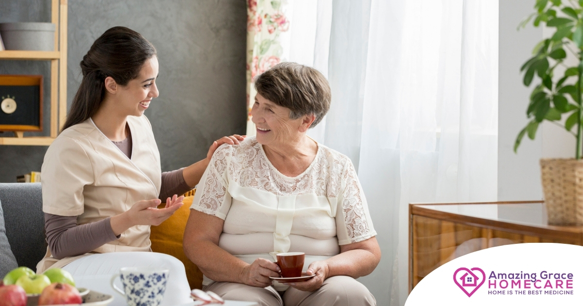 Smiling caregiver with her hand on an older woman’s shoulder as they share tea on the couch, showing home care vs assisted living
